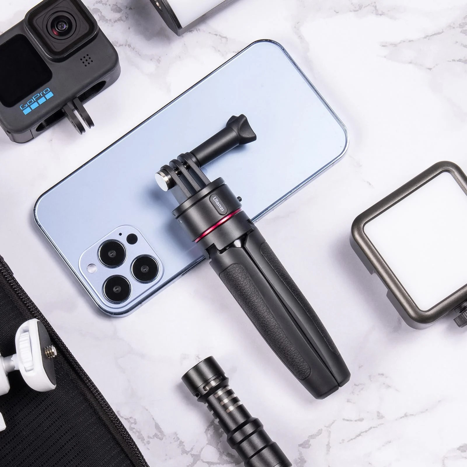 Un mini treppiede nero poggia su uno smartphone azzurro, circondato da GoPro, microfono e luce su superficie di marmo.