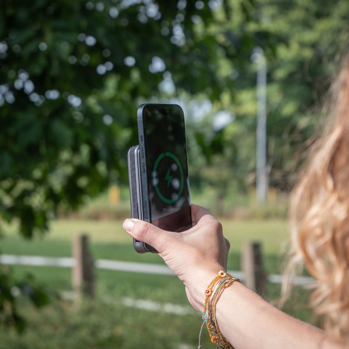 Eine Person hält ein Smartphone mit einer magnetischen Powerbank, die einen grünen Ladekreis zeigt, vor einem Park.
