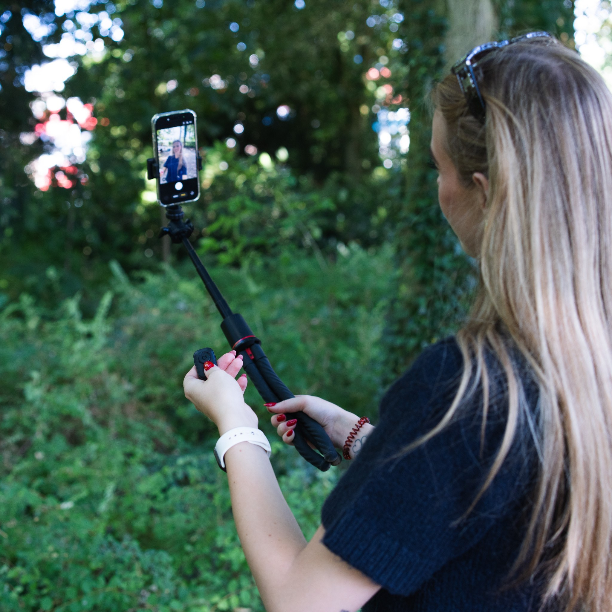 Une femme blonde dans une forêt tient un smartphone sur un trépied pour un selfie.