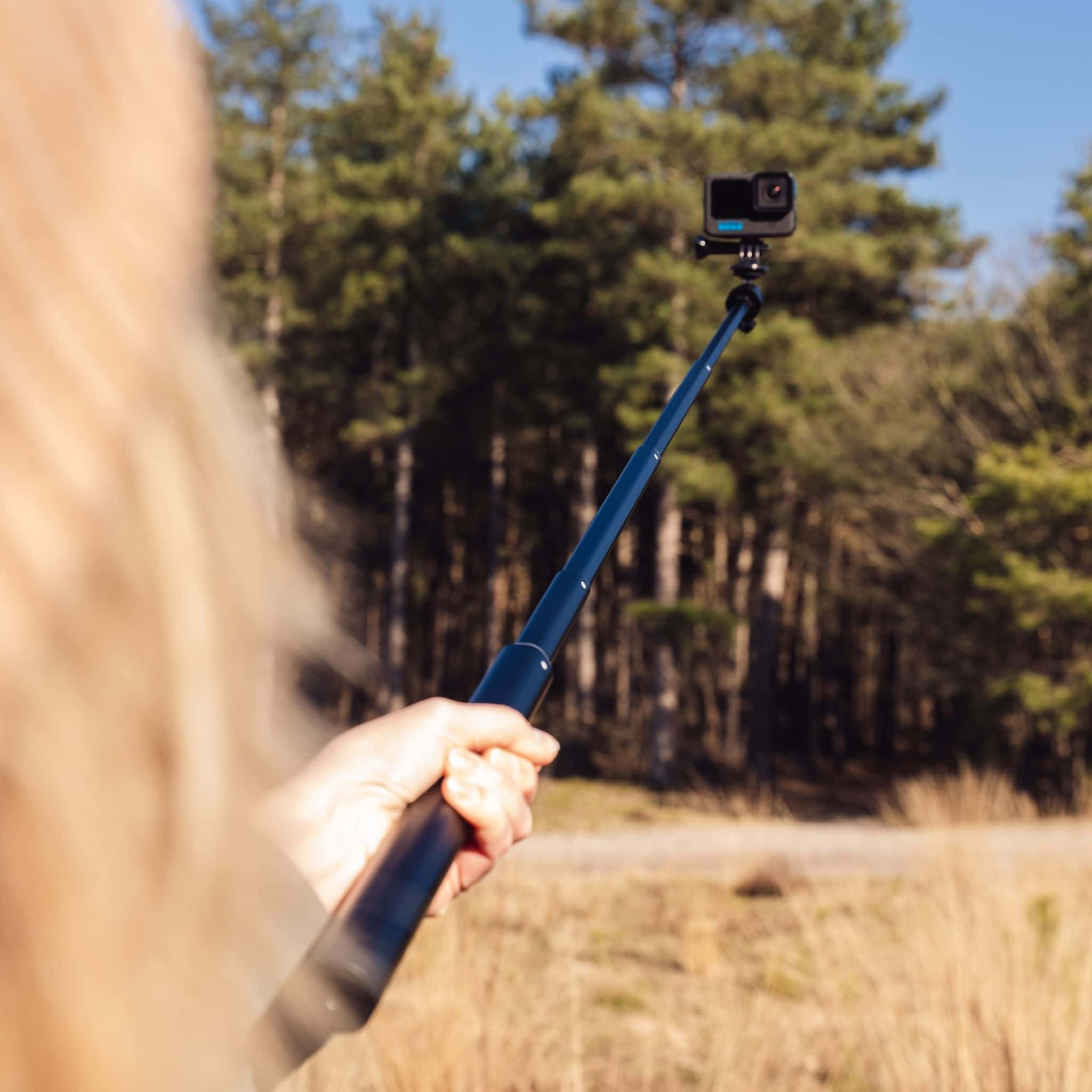 Une femme blonde tenant un selfie stick avec une caméra d'action dans une forêt.