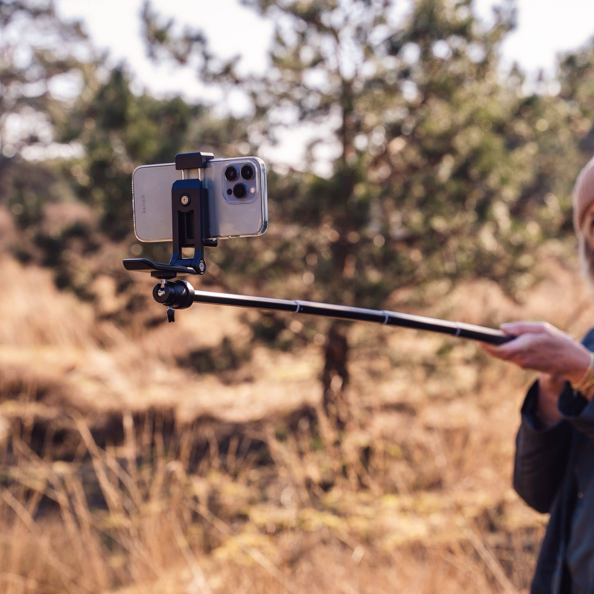 Une personne tient une perche à selfie avec un smartphone à l'extérieur devant une forêt floue.