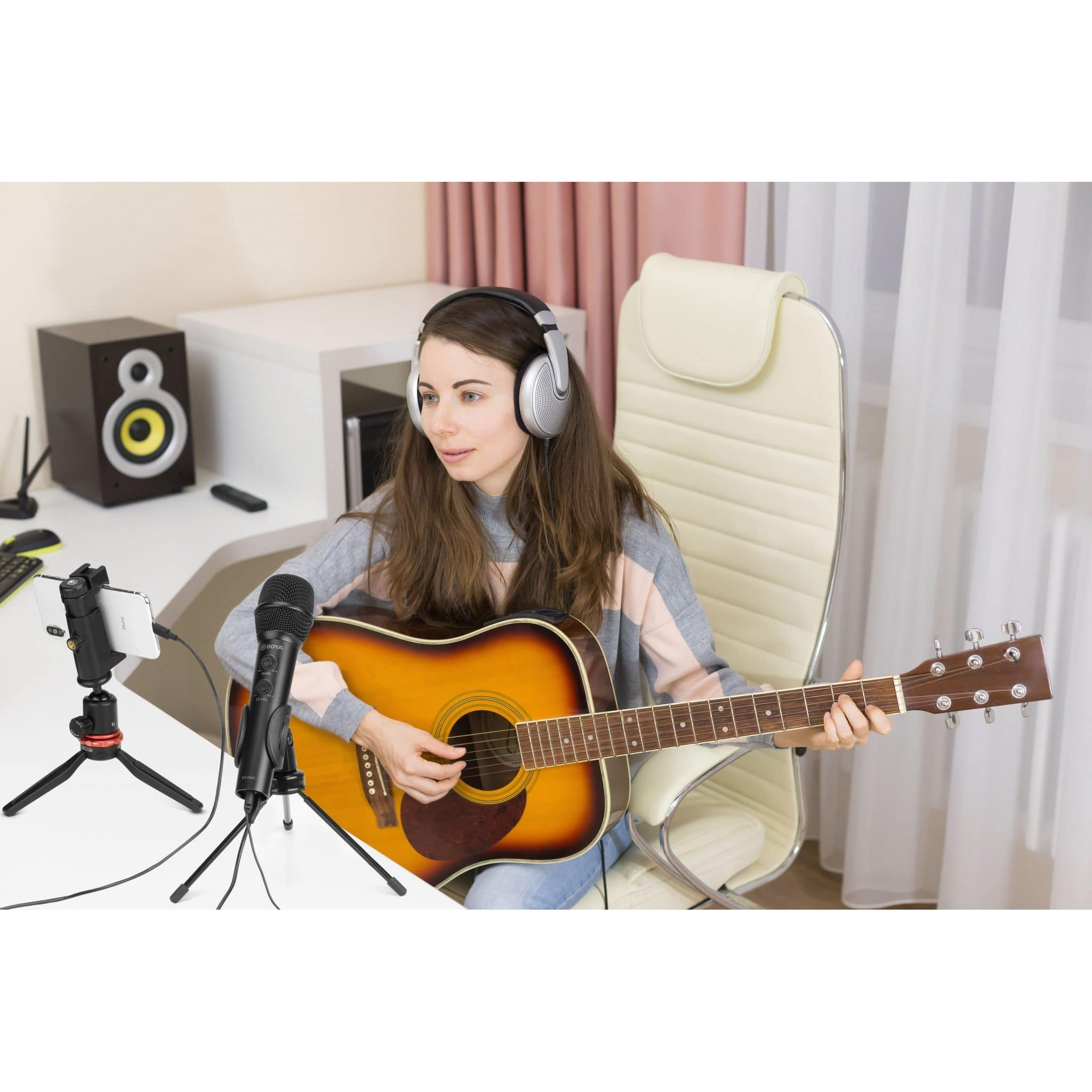 Frau sitzt in Kopfhörern auf einem Stuhl, spielt Gitarre vor einem Mikrofon und Smartphone.
