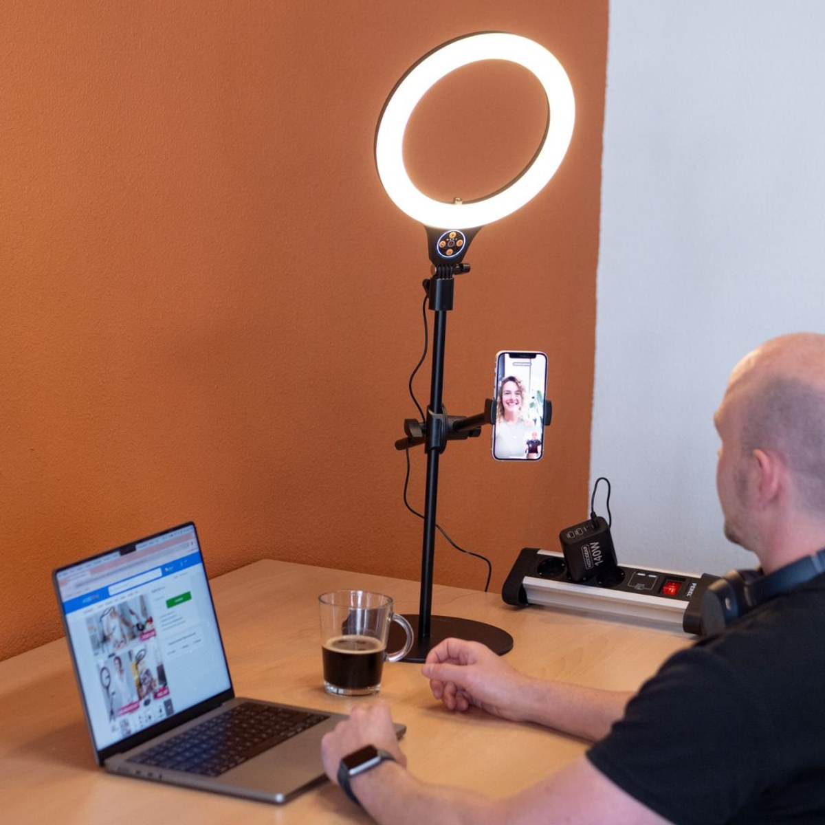 Een man zit aan een houten bureau met laptop, ringlamp en smartphone.