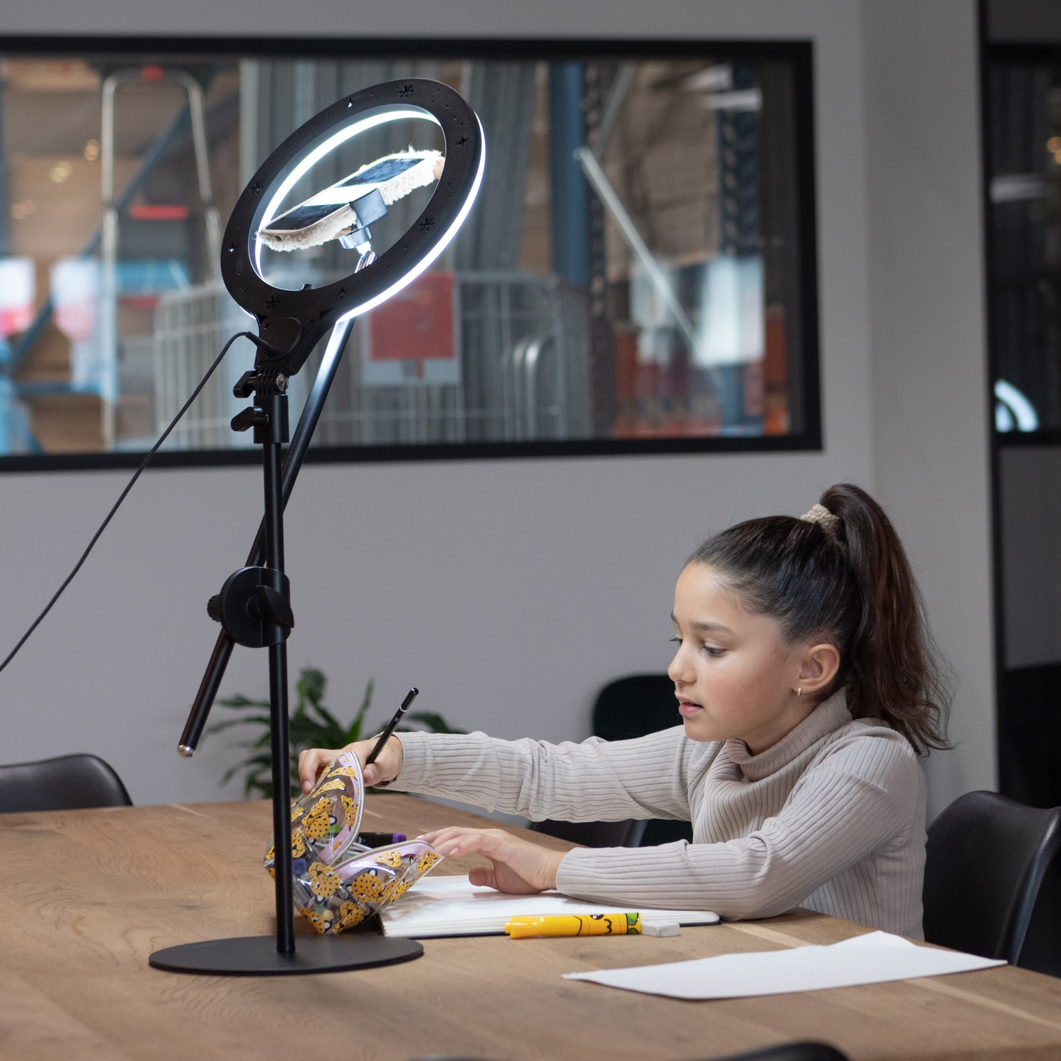 Een jong meisje met een paardenstaart zit aan een houten tafel en opent een etui voor een verlichte ringlamp.