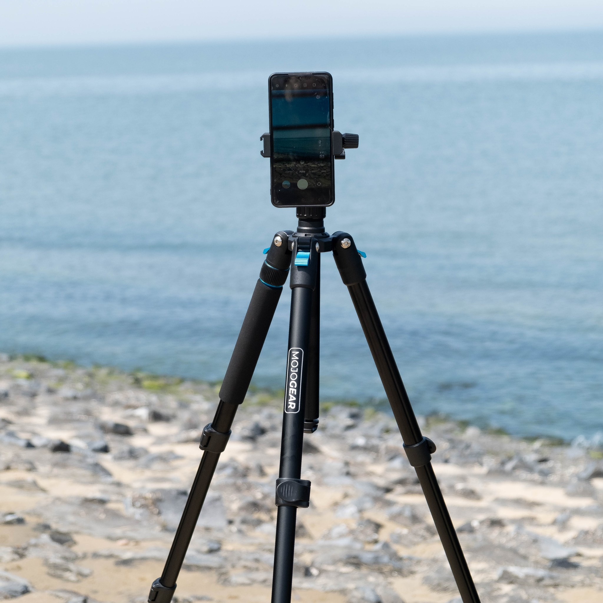 Un trépied noir avec un smartphone sur une plage rocheuse.