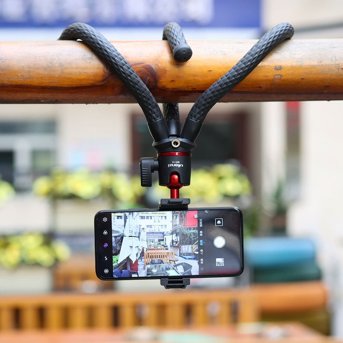Un treppiede flessibile con smartphone è appeso a testa in giù a un palo di legno in un ambiente esterno sfocato.