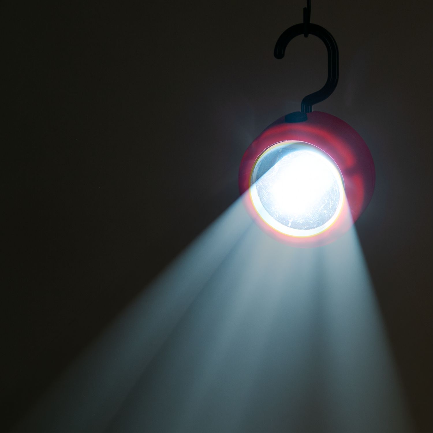 Eine rote Hängearbeitsleuchte mit Haken strahlt im Dunkeln einen hellen weißen Lichtkegel diagonal nach unten ab.