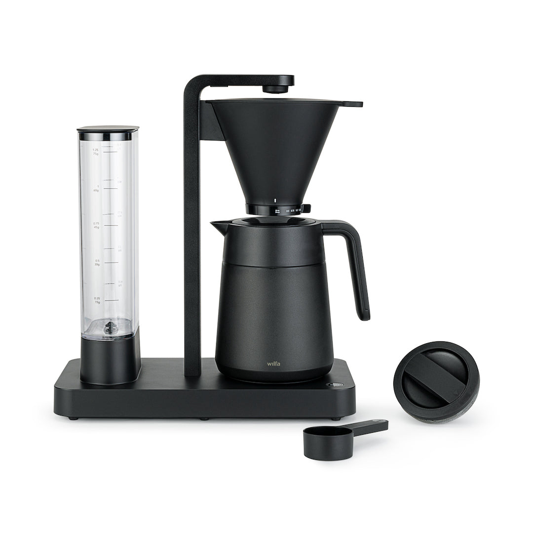 Moderna macchina da caffè a filtro nera Wilfa con serbatoio trasparente, caraffa, misurino e coperchio su fondo bianco.