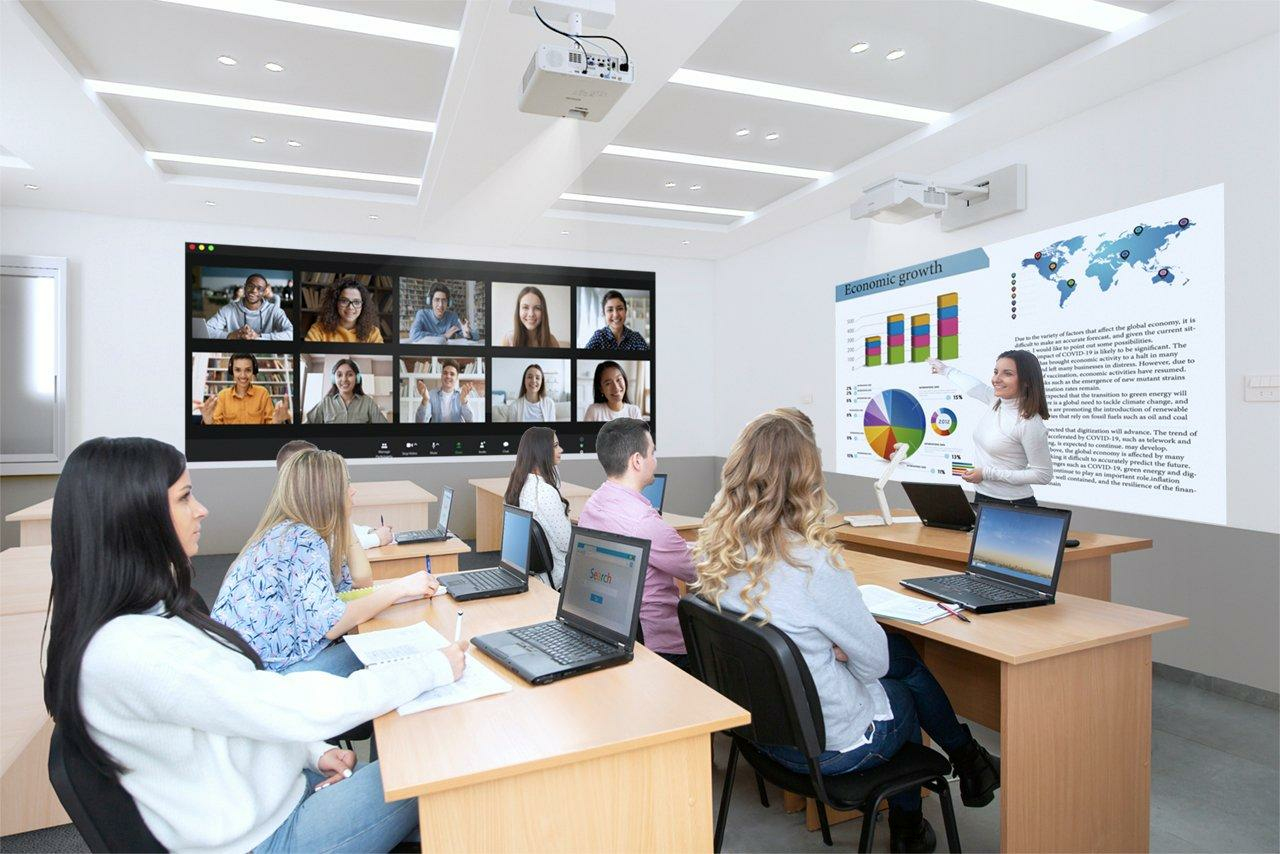 Eine Lehrerin präsentiert vor Studenten, die an Laptops sitzen, während ein Videoanruf auf einer Leinwand läuft.