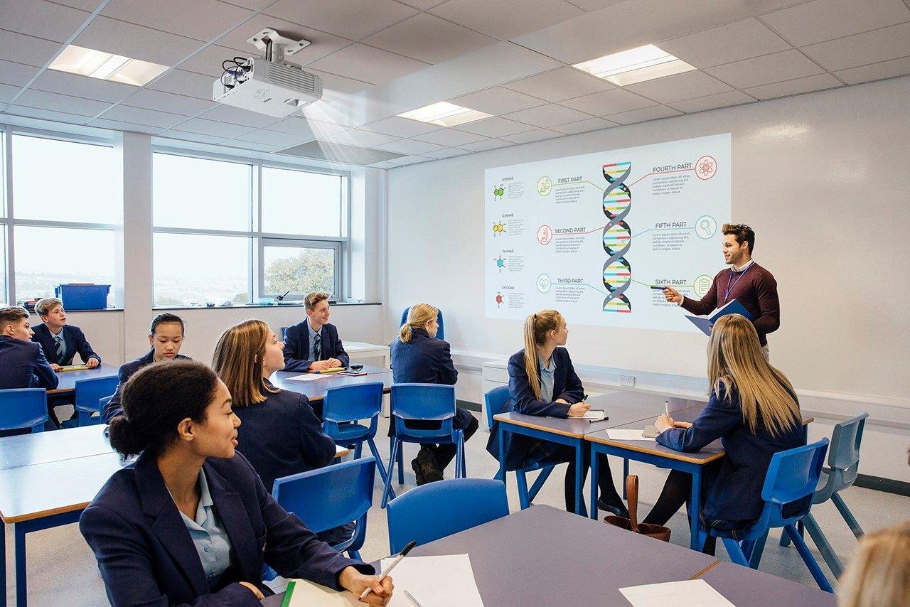 Ein Lehrer steht vor einer Leinwand mit Schülern in einem hellen Klassenzimmer.