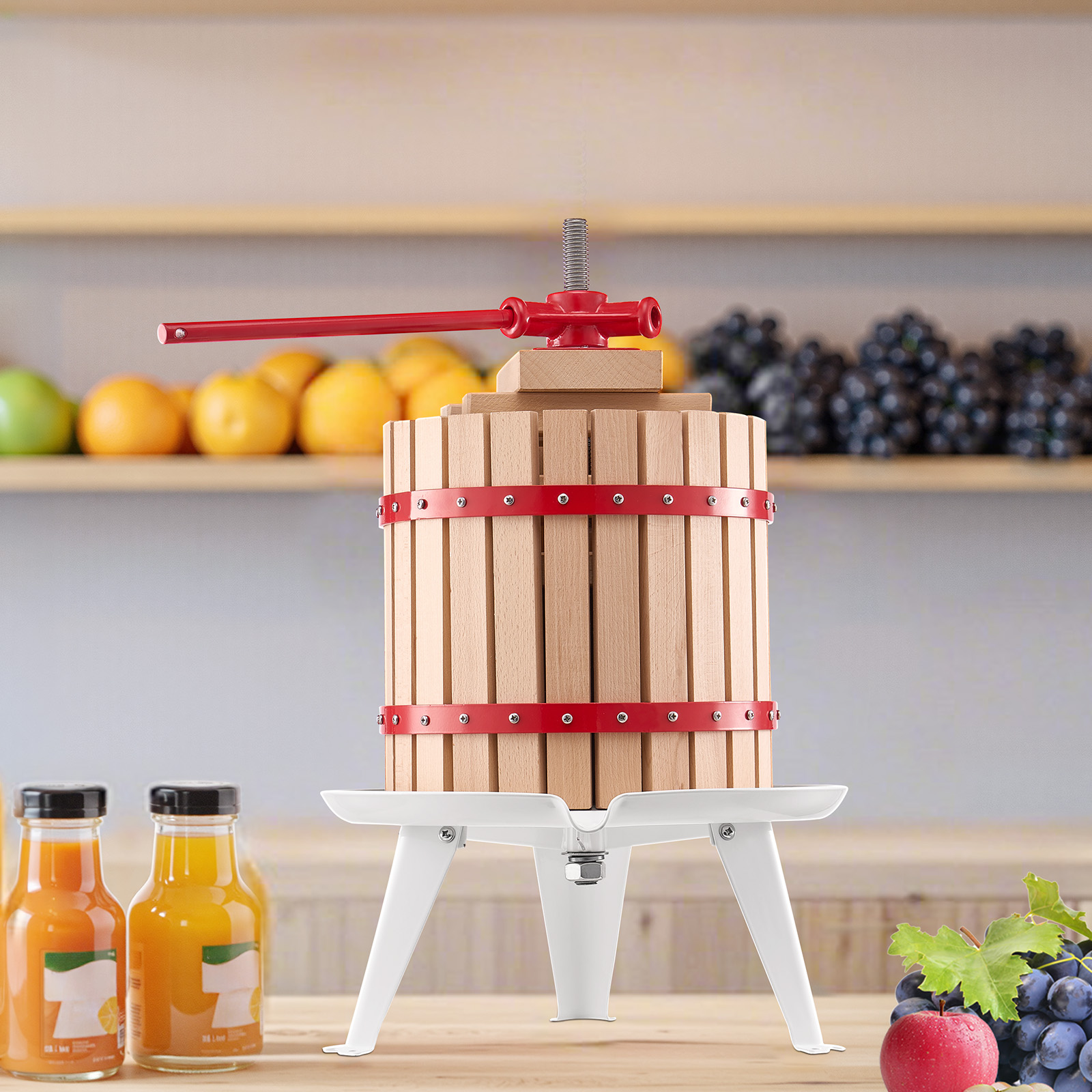 Una prensa de fruta de madera con bandas metálicas rojas se encuentra en una mesa, rodeada de botellas de jugo y fruta fresca.