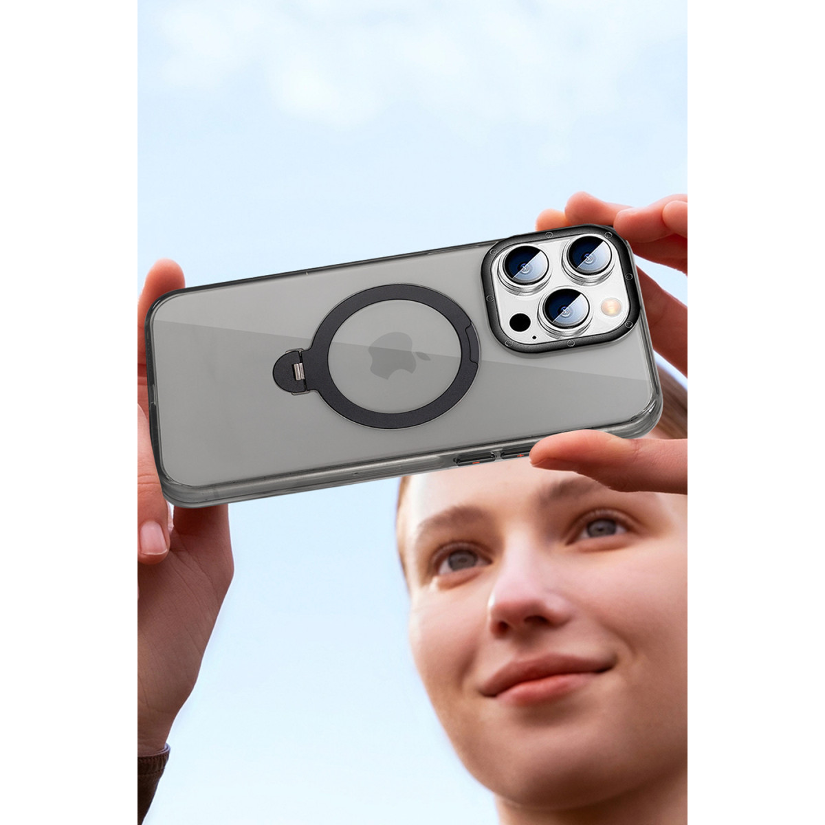 Bir kişi açık havada, siyah MagSafe halkalı şeffaf kılıftaki akıllı telefonu yukarıda tutuyor.
