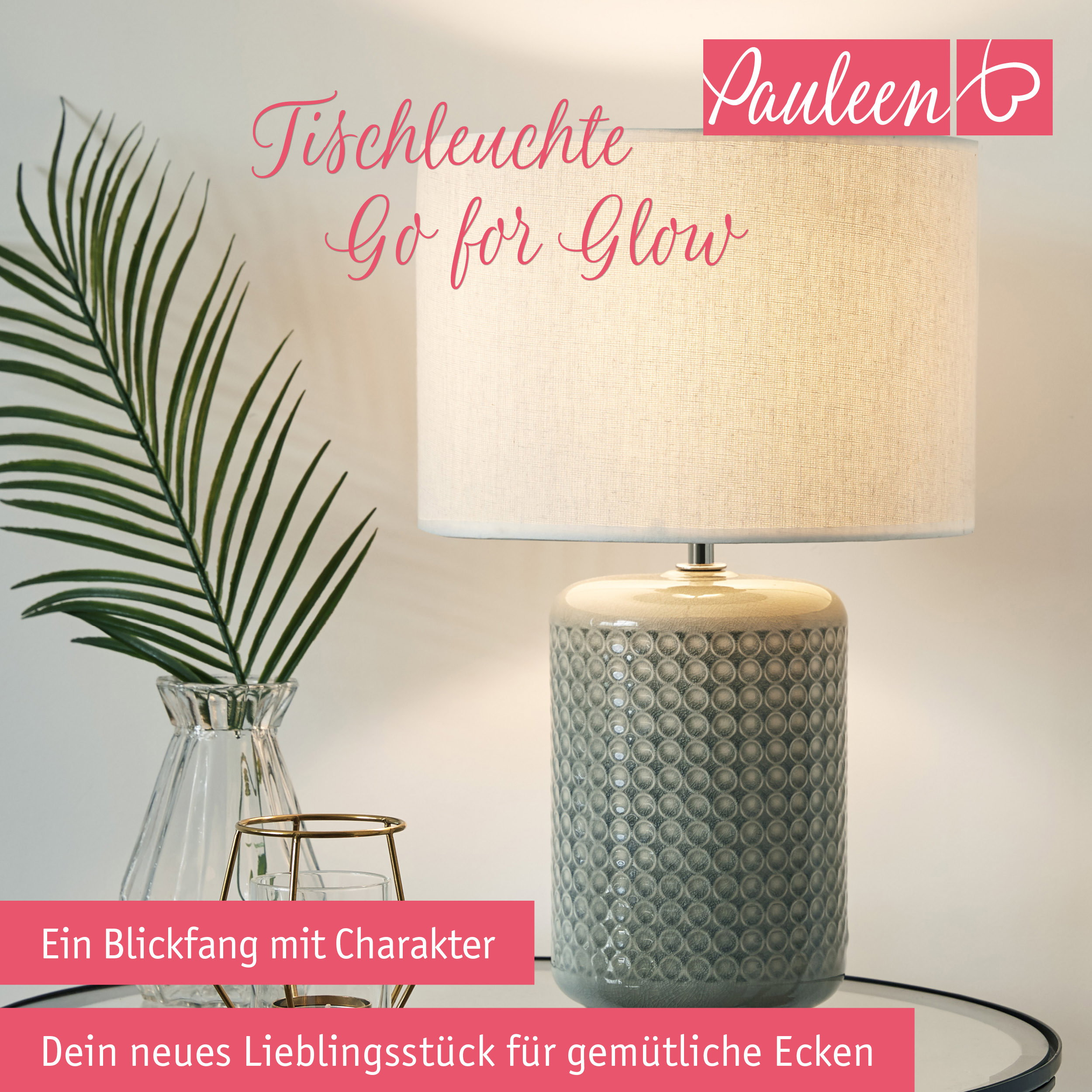 Beigefarbene Tischleuchte mit Reliefmuster, Glasvase mit Palmblatt und Kerzenhalter auf einem Tisch.