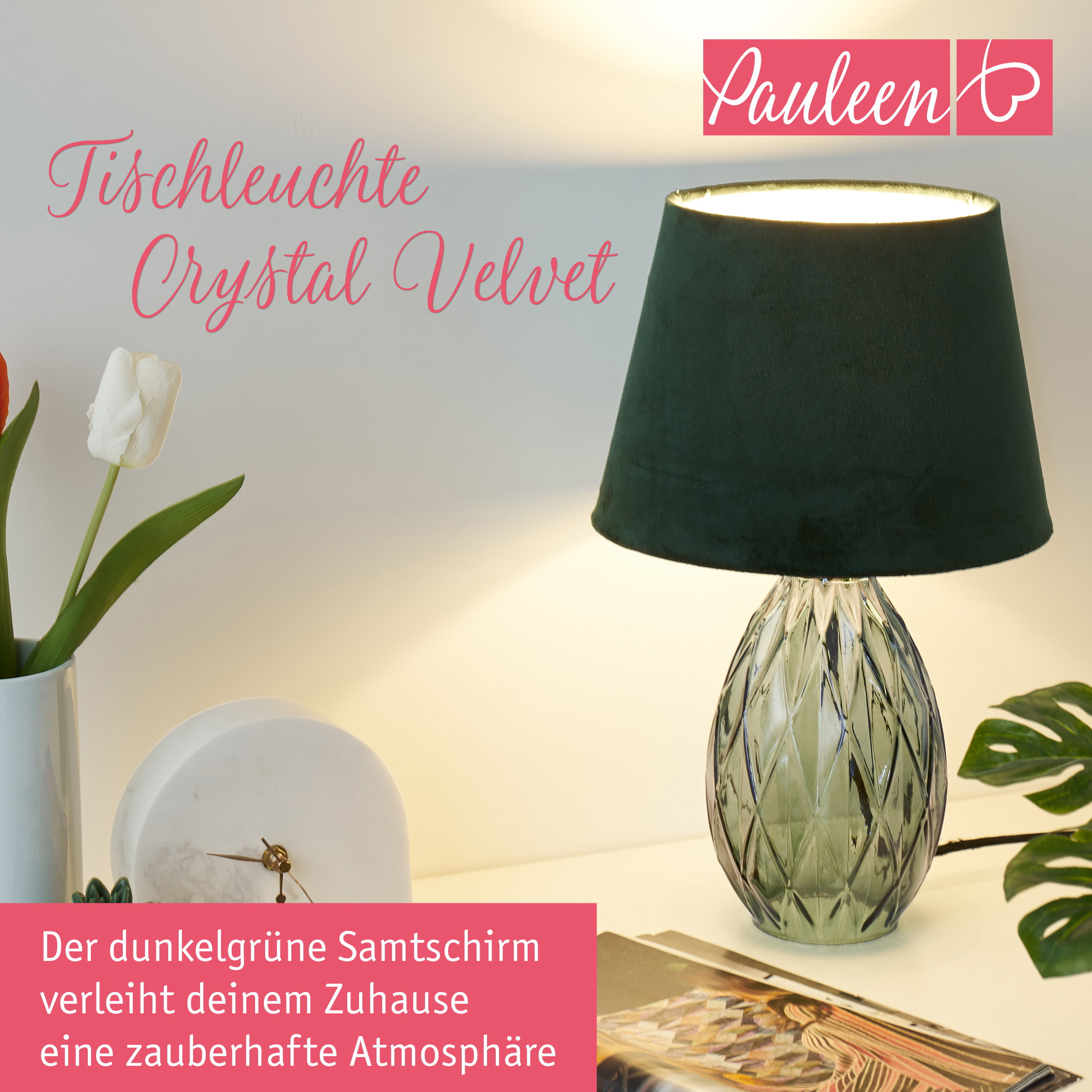 Eine grüne Tischleuchte mit Samtschirm und Facettenfuß, dekoriert mit einer Tulpe und einer Uhr auf einem Tisch.