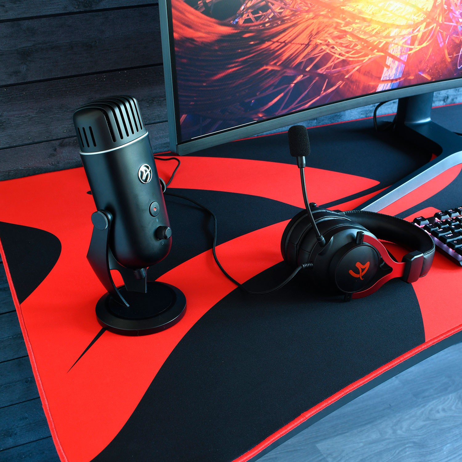 Micrófono de pie negro y auriculares para juegos en un escritorio rojo y negro ante un monitor.