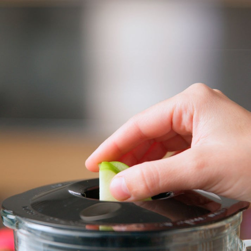 Una mano inserisce una fetta di mela verde nel coperchio di un frullatore con sfondo sfocato.