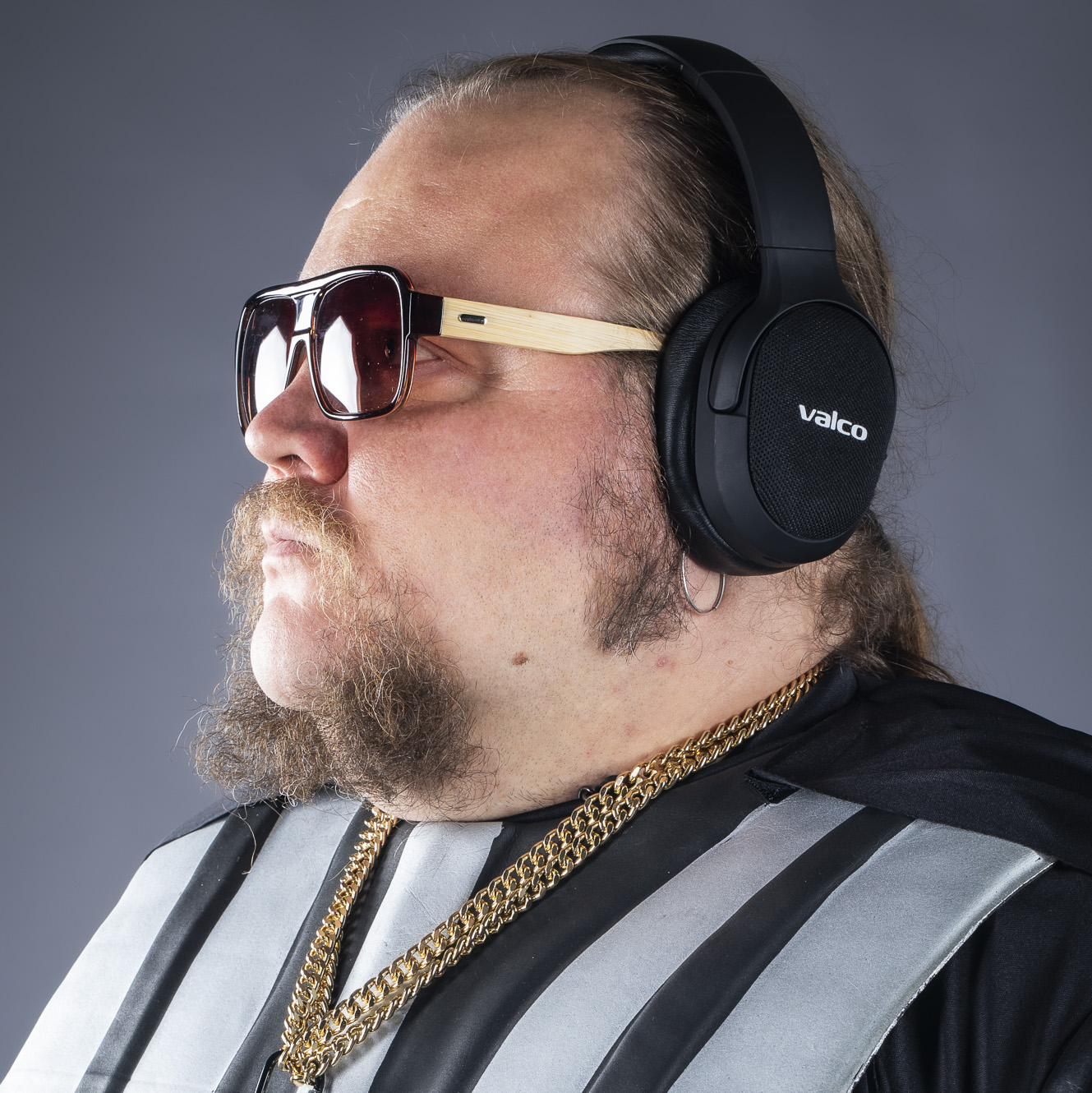 Ein Mann mit Bart, Sonnenbrille, Kopfhörern und goldenen Ketten vor grauem Hintergrund.