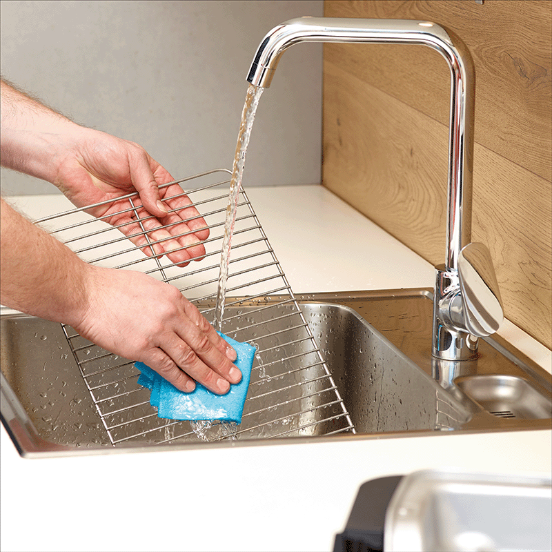 Person wäscht ein Ofengitter mit einem blauen Tuch unter fließendem Wasser im Küchenwaschbecken.