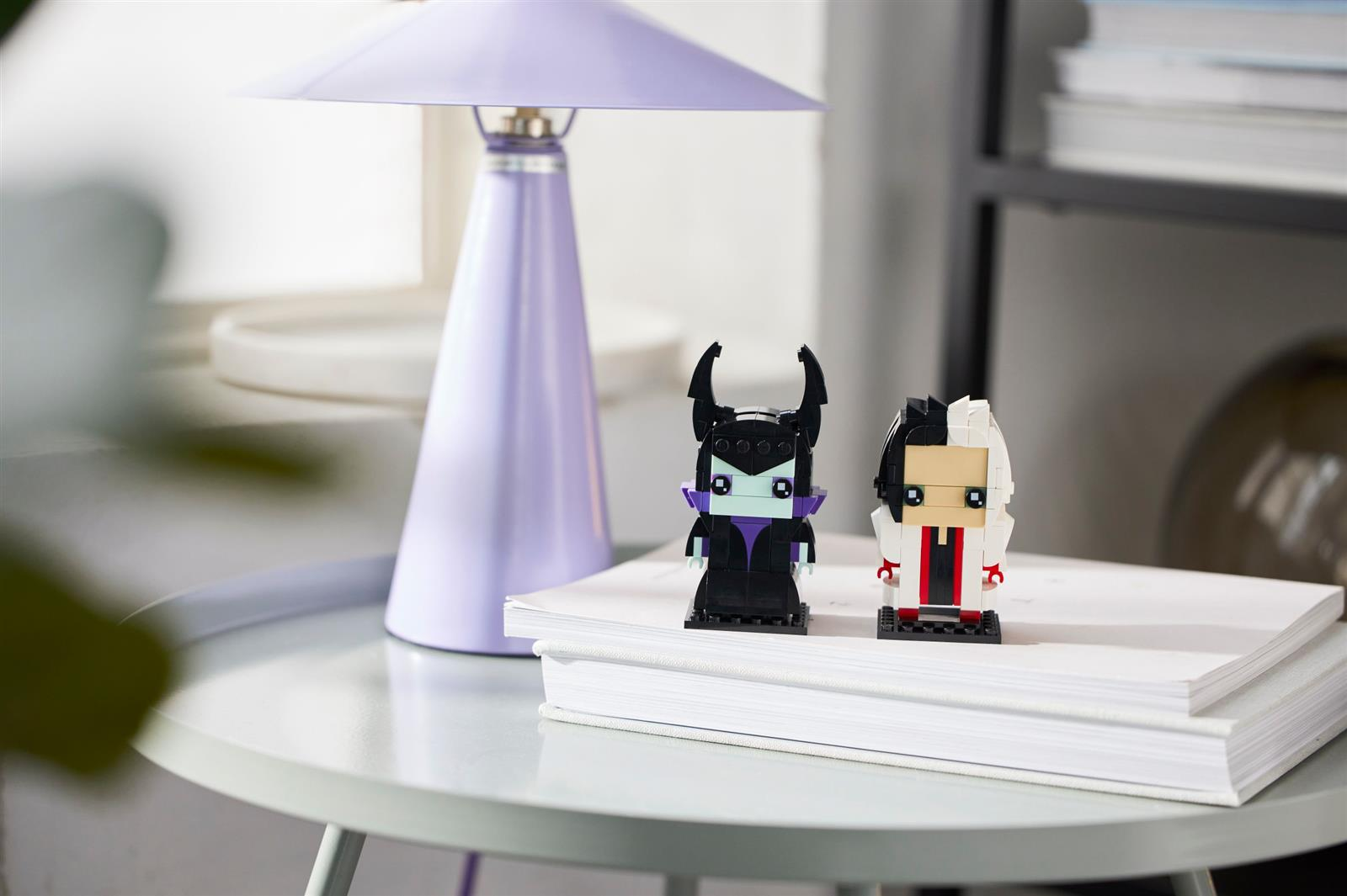Zwei LEGO Figuren stehen auf einem Stapel weißer Bücher neben einer lilafarbenen Lampe auf einem Tisch.