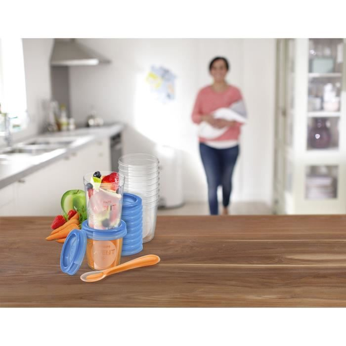 Babynahrungsbehälter mit Obst, Löffel und Bechern auf einem Holztisch, im Hintergrund eine Mutter mit Baby.