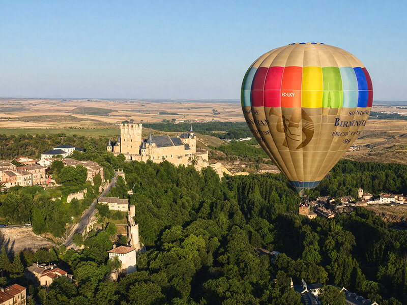 Colorido globo aerostático vuela sobre un valle verde con un castillo histórico y una ciudad al fondo.