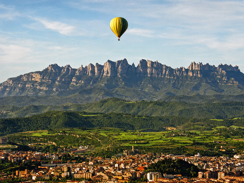 Un globo aerostático amarillo y verde vuela sobre un pueblo, colinas verdes y montañas escarpadas.