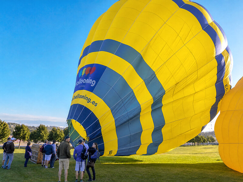 Un gran globo aerostático amarillo y azul se prepara en un campo verde con personas cerca.