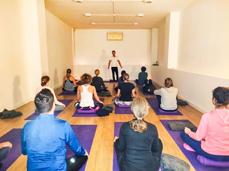 Un grupo de yoga se sienta en esterillas moradas en una habitación luminosa, mirando al instructor.