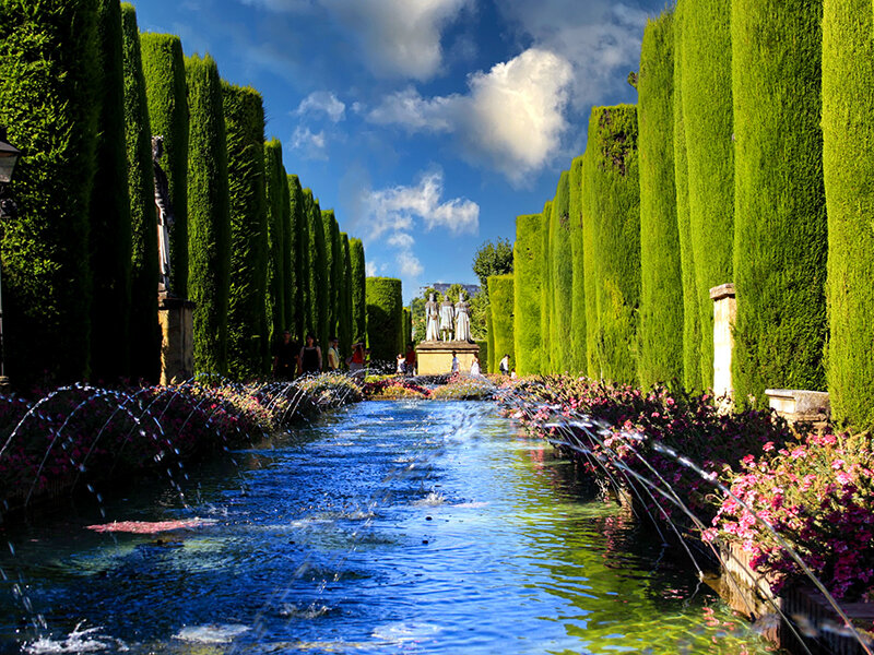 Larga fuente de agua en un jardín flanqueado por altos árboles verdes.