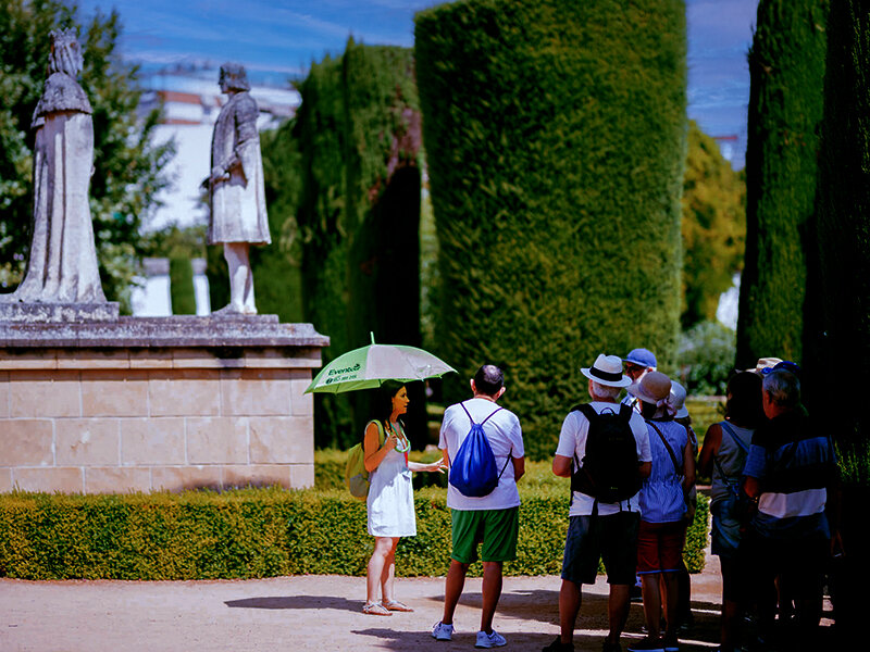 Un guía turístico con paraguas verde explica dos estatuas a un grupo de turistas en el jardín.