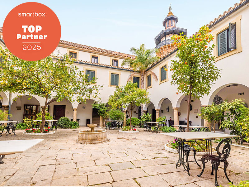 Soleado patio de un edificio histórico con árboles de cítricos, mesas y una fuente.