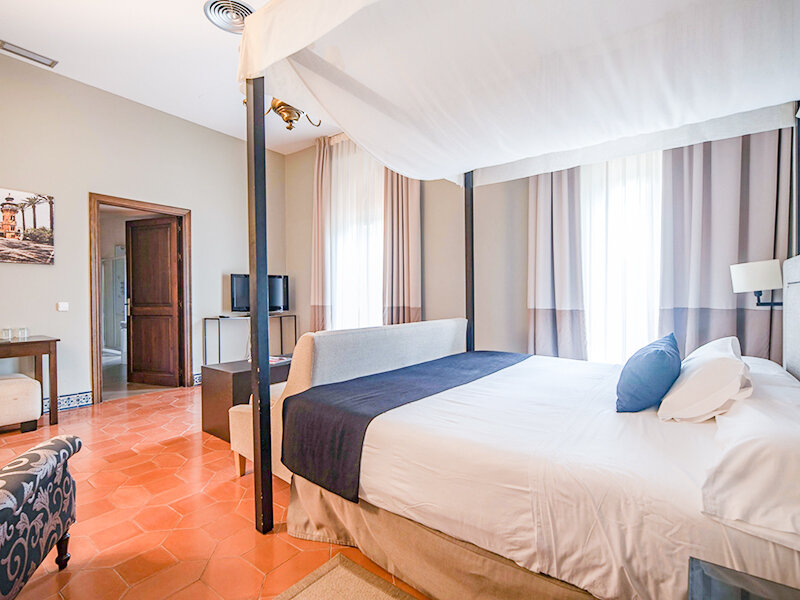 Dormitorio con cama con dosel, paredes blancas, suelo de terracota y puerta de madera abierta.
