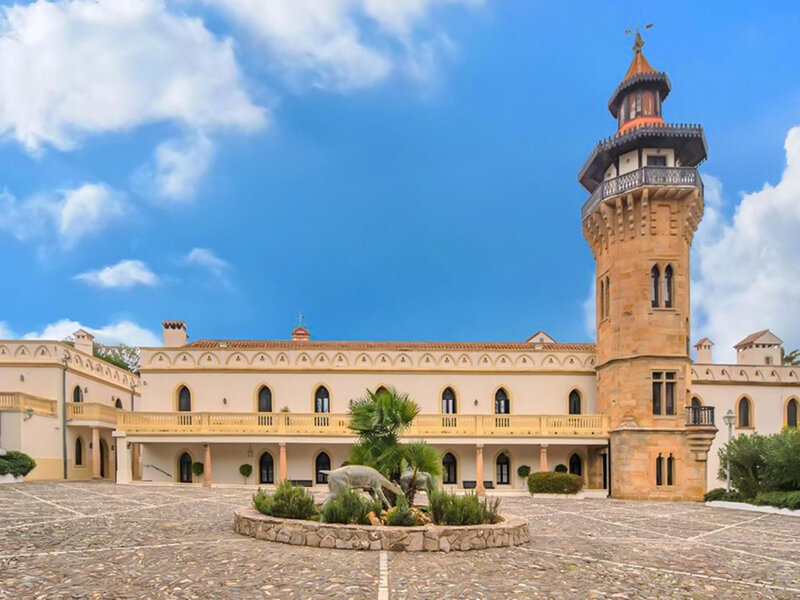 Un palacio blanco con una torre de piedra alta, una terraza con balcón y un patio de jardín de piedra.