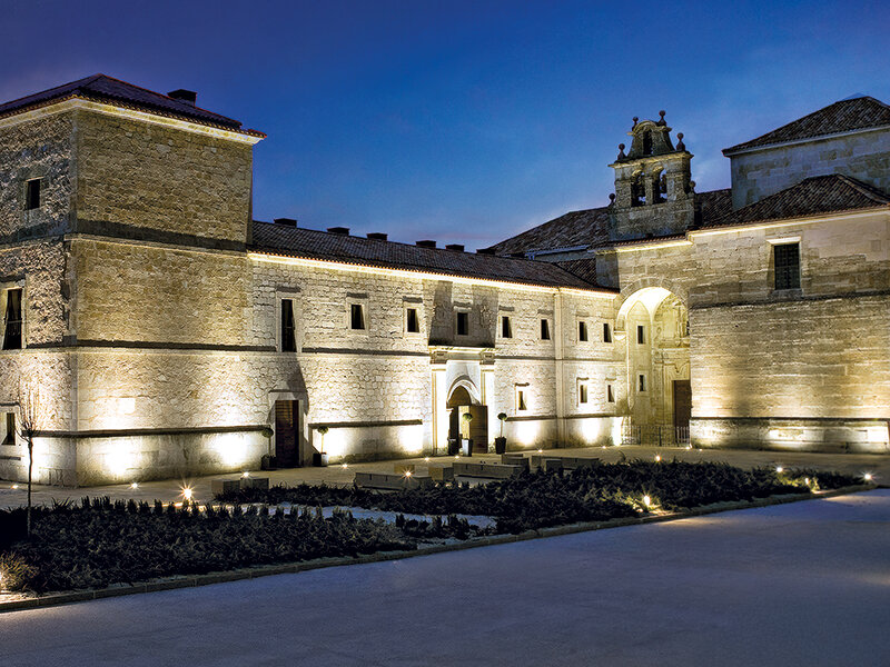 Edificio histórico de piedra iluminado al atardecer con un patio y jardines en primer plano.