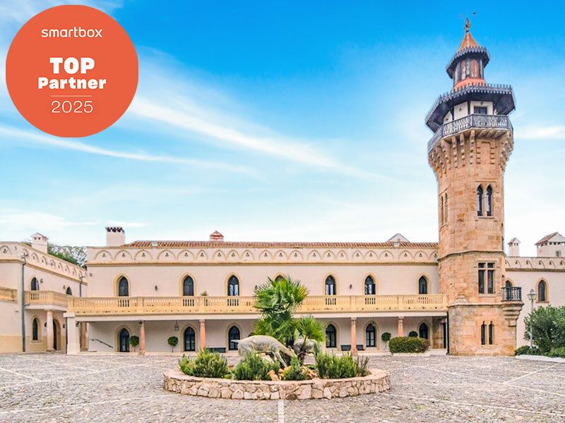 Un castillo histórico con una torre alta y un patio de piedra en primer plano bajo un cielo azul.
