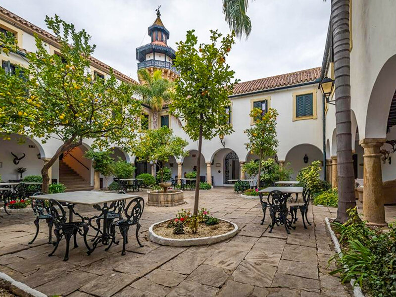 Un patio soleado con suelo de piedra, cítricos, mesas, sillas y una torre decorativa alta al fondo.