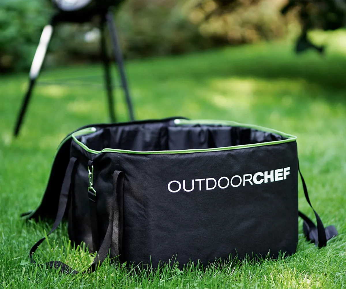 Yeşil çimler üzerinde açık siyah Outdoorchef çanta, arka planda bulanık bir mangal var.
