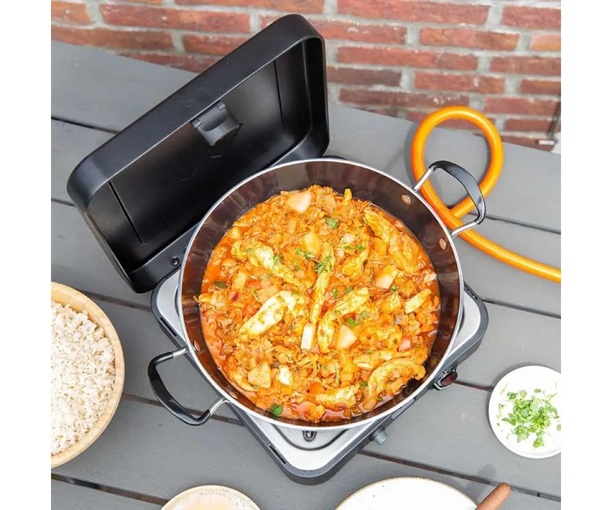Ahşap masa üzerinde gazlı ocakta pişen tavuklu ve sebzeli bir paella tavası.