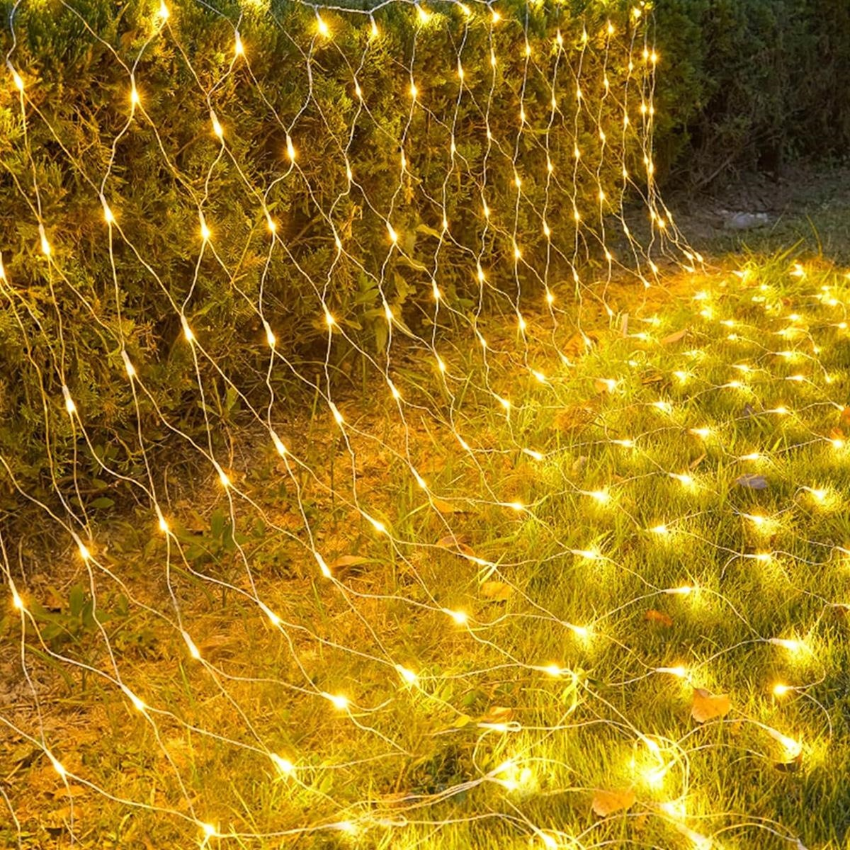Helle, warmgelbe Lichterketten über grünen Büschen und Gras, die eine gemütliche Abendstimmung erzeugen.