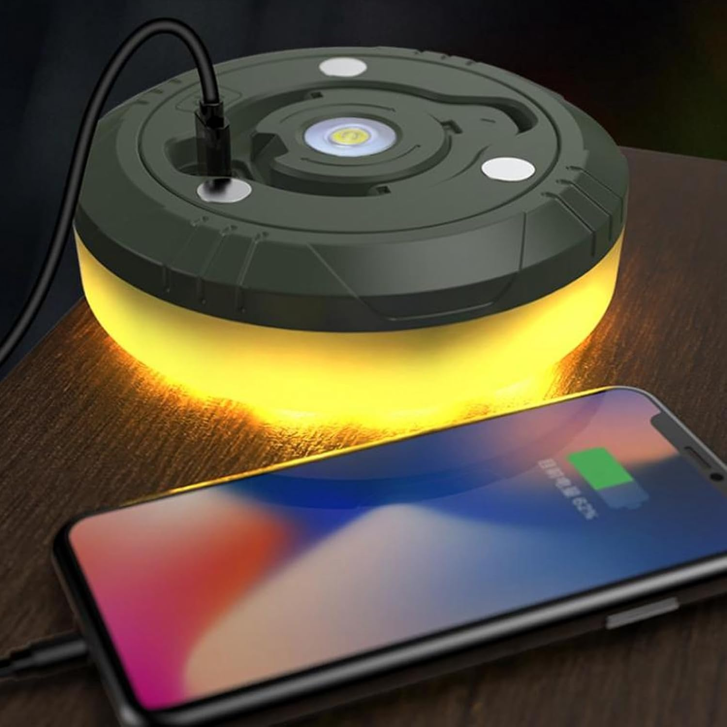 Eine runde, grüne Campinglaterne leuchtet gelb auf einem Holztisch neben einem Smartphone.