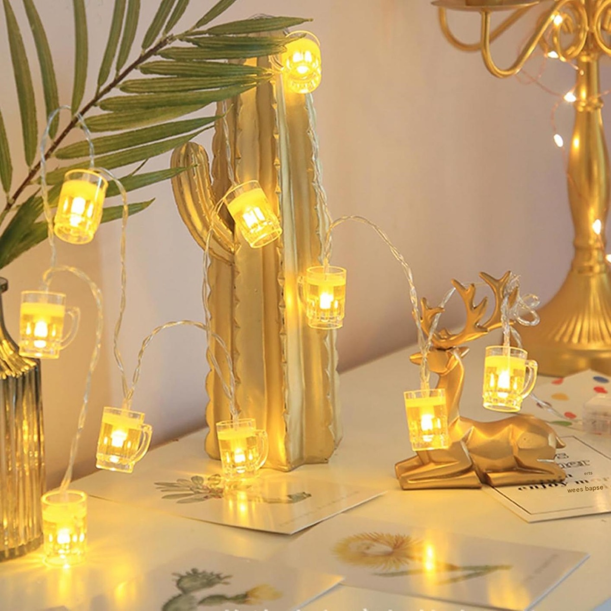 Eine Lichterkette mit leuchtenden Bierkrug-Lichtern liegt auf einem goldenen Kaktus und einer Rentierfigur.