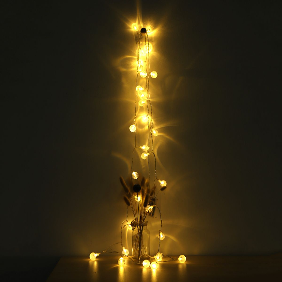 Eine Lichterkette mit warmen, leuchtenden Lichtern fließt an einer dunklen Wand hinter einer Glasvase mit getrockneten Blumen herunter.