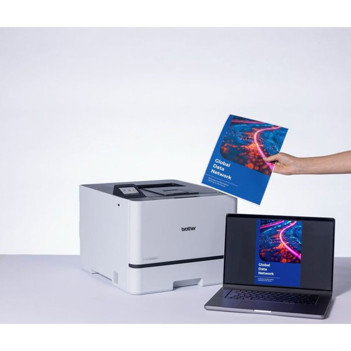 Ein weißer Brother-Drucker, ein Laptop mit Dokument auf dem Bildschirm und eine Hand, die ein ausgedrucktes Dokument hält.