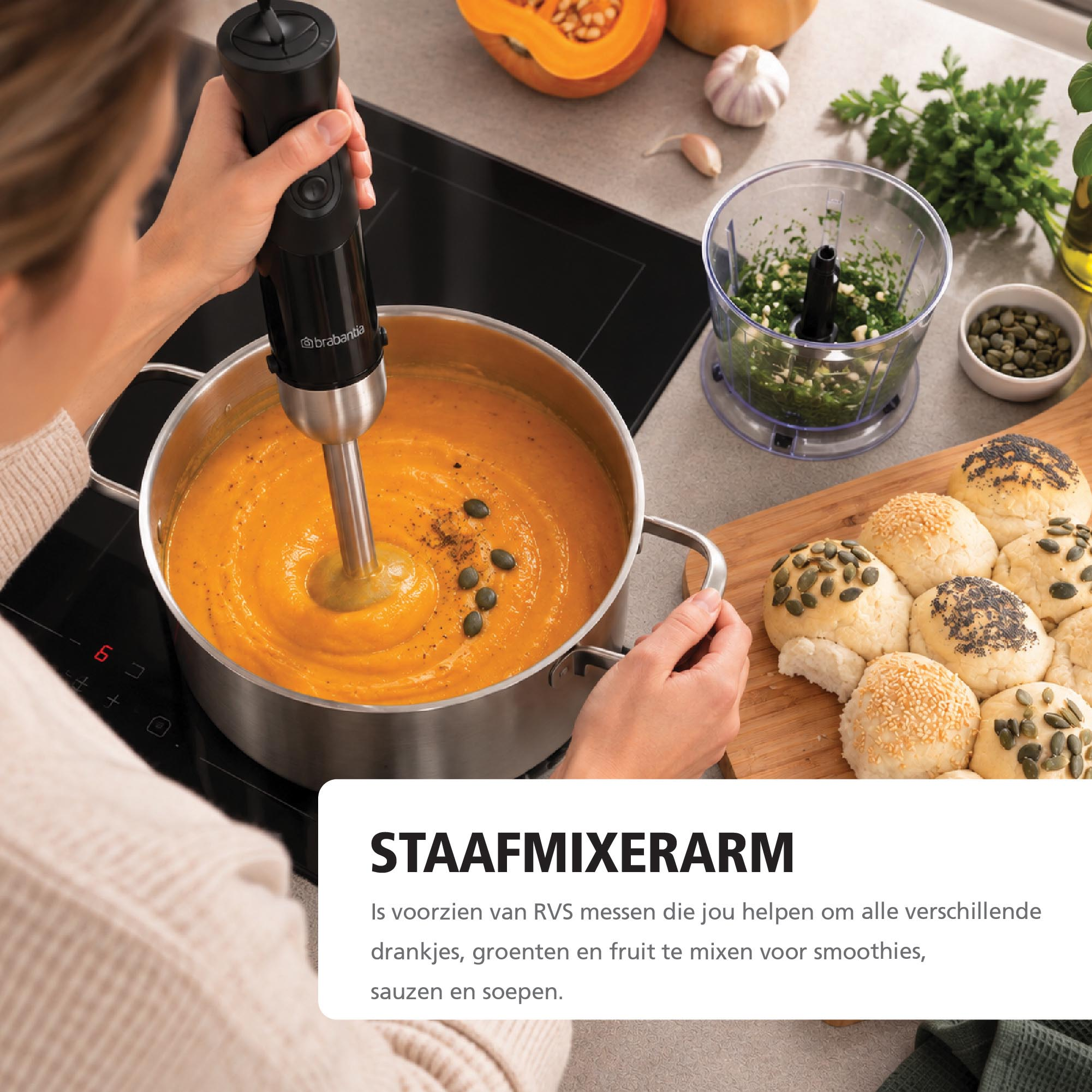 Woman blends orange soup with black hand blender in metal pot near rolls.