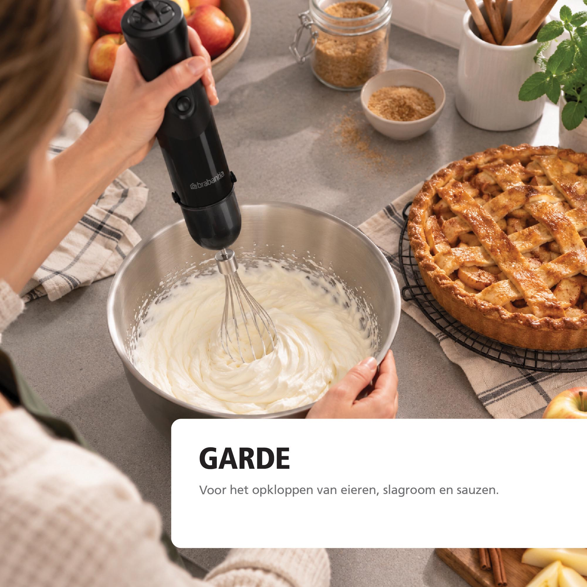 Person mixes cream in metal bowl with black hand blender. An apple pie and baking ingredients are on the counter.