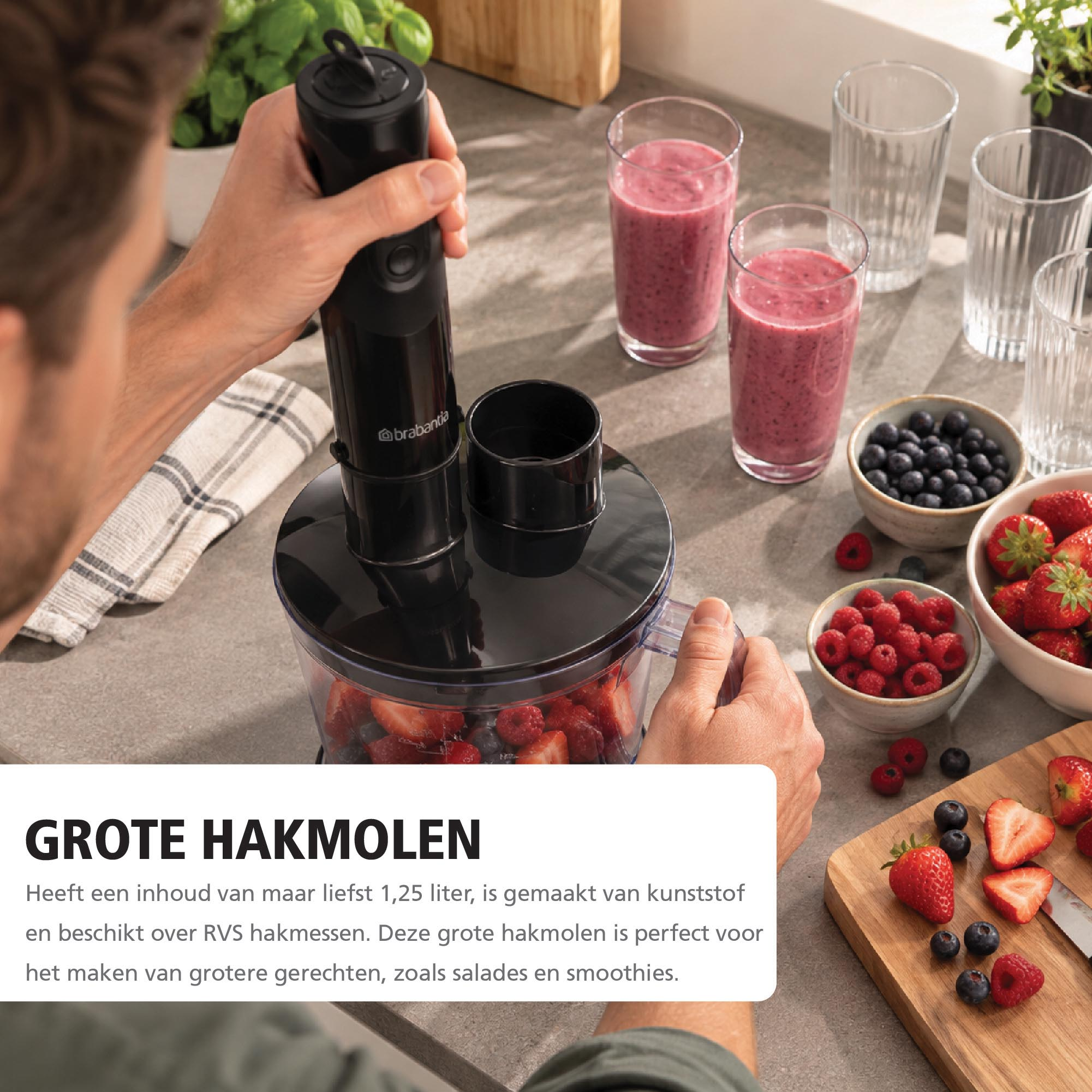 A man uses a black hand blender over a bowl, surrounded by berries, smoothies, and a cutting board on a counter.