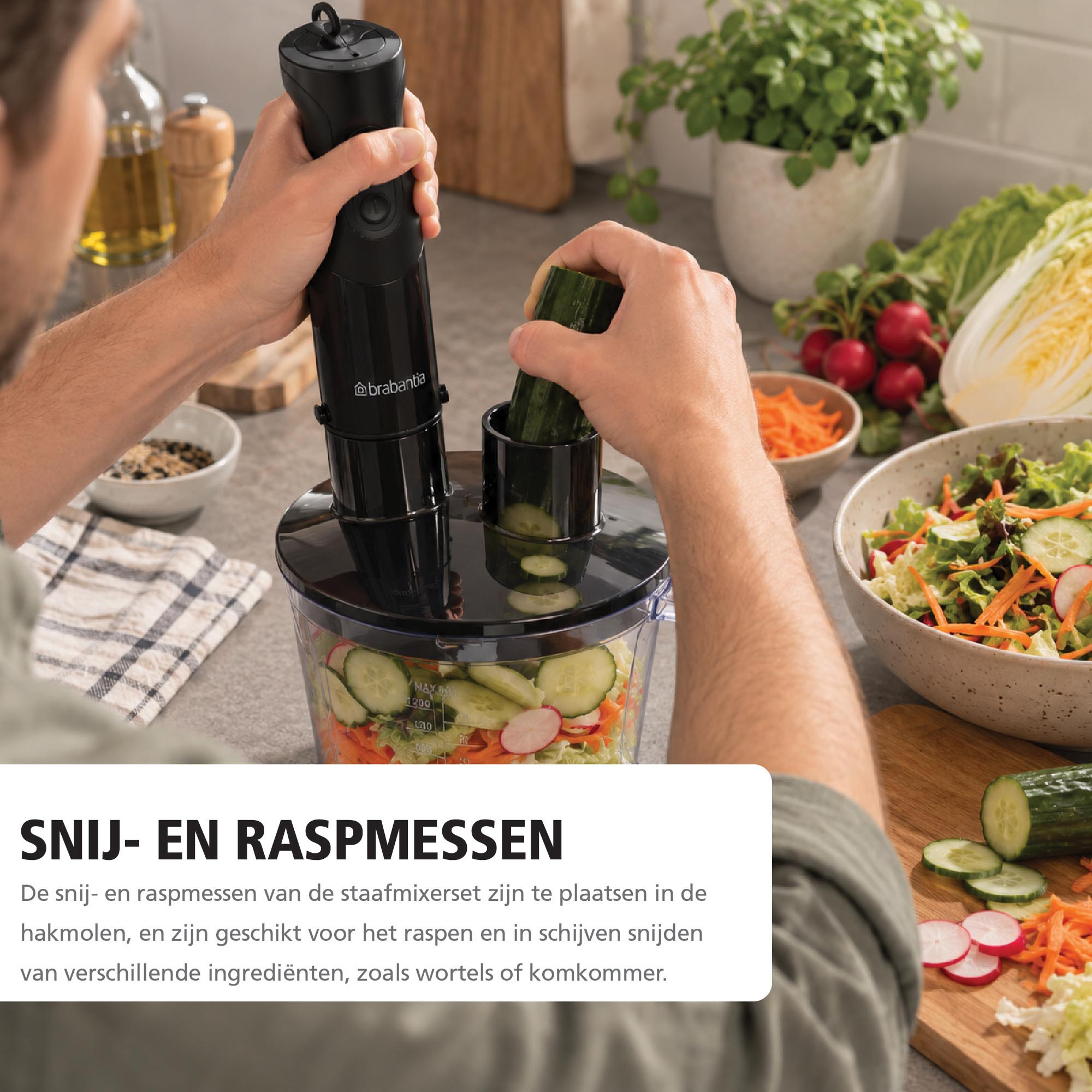 A person uses a black food processor with a cucumber, surrounded by chopped salad, vegetables, and kitchen items.