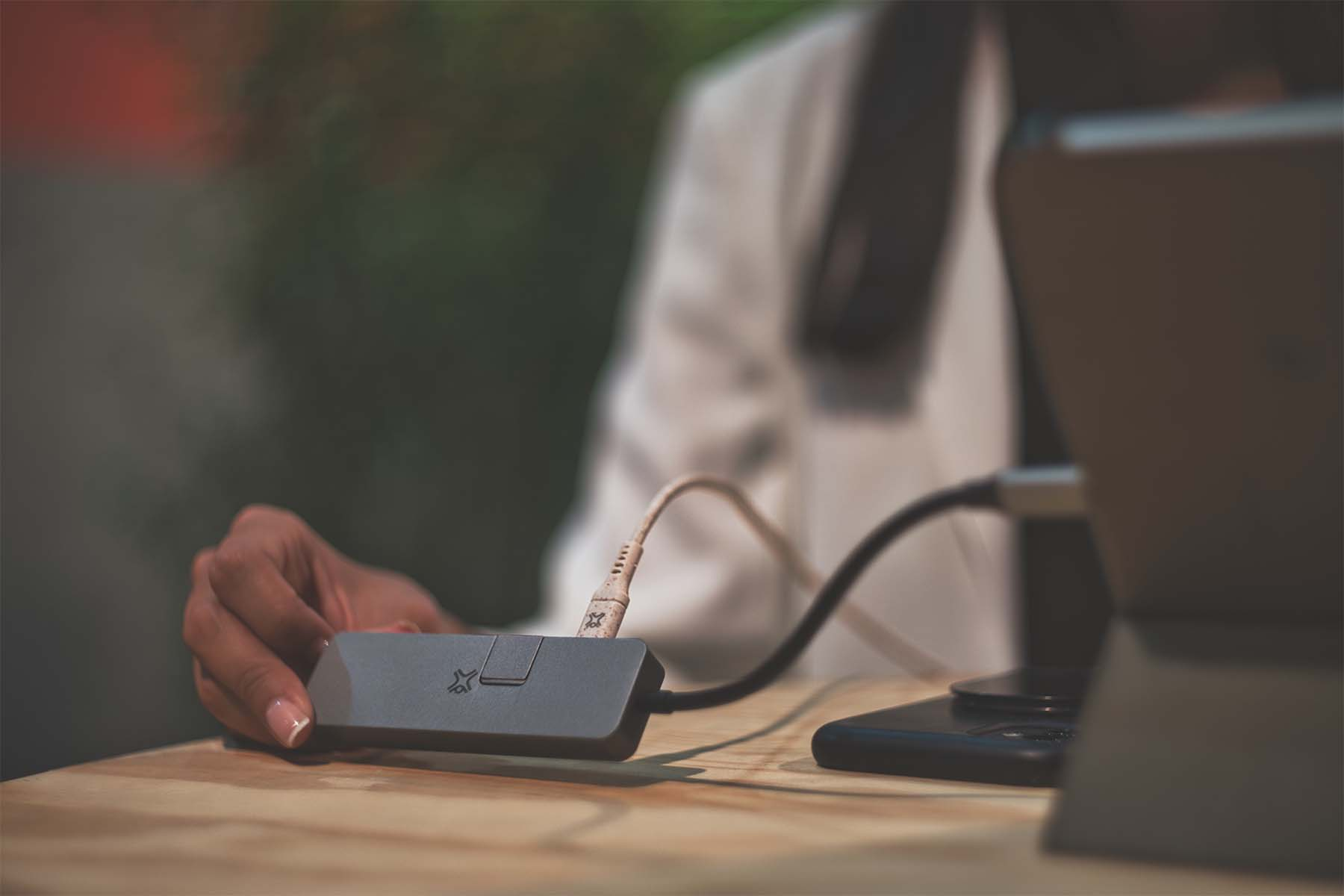 Una mano tiene un hub USB-C grigio su un tavolo di legno, con i cavi collegati.
