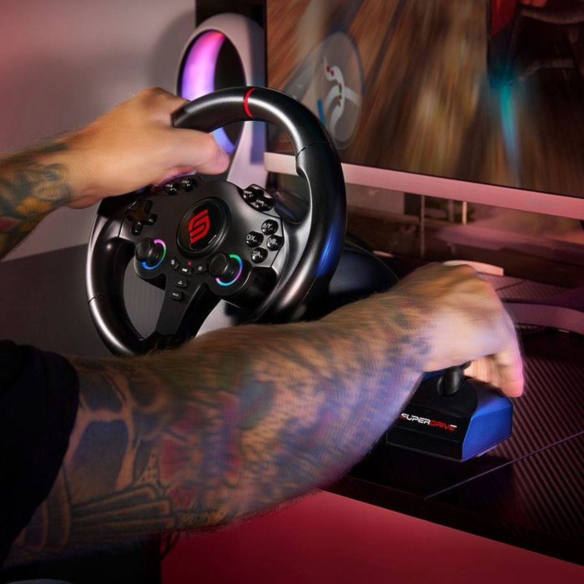 Tätowierte Hände bedienen ein schwarzes Gaming-Lenkrad und einen Schalthebel vor einem Monitor.