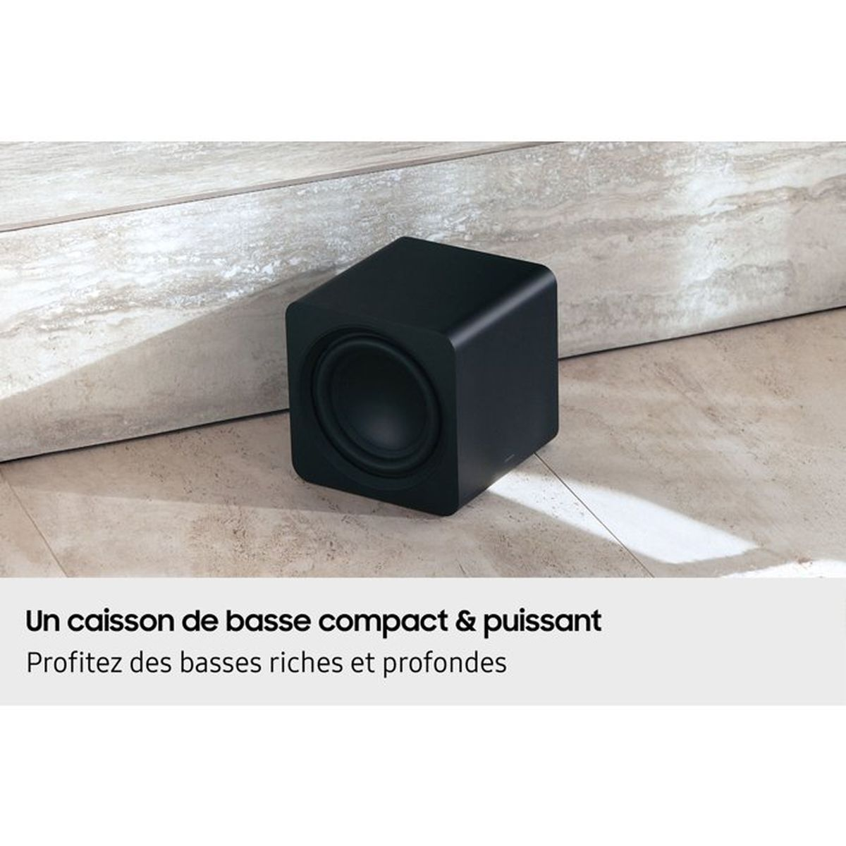 Ein kompakter, schwarzer Subwoofer steht auf einem gefliesten Boden vor einer hellen Wand.